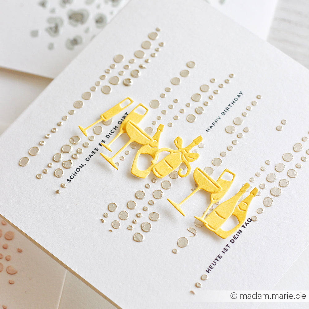 Weiße Geburtstagskarte mit goldenen Weinglas-Ausschnitten, geprägten Punkten im Schablonen-Punktmuster sowie deutschen und englischen Grußtexten. Perfekt für Schablonen-Papierprojekte oder zur Verwendung mit wiederverwendbaren Schablonen, um Sie für Ihre nächste kreative Feier zu inspirieren.