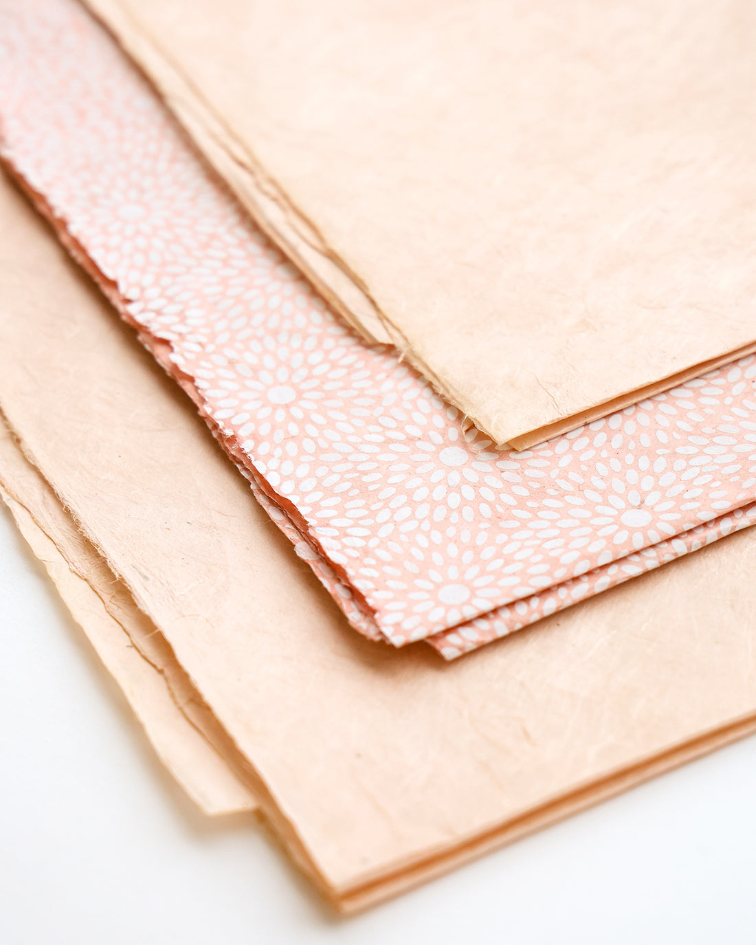 Drei Blätter aus strukturiertem, pastellrosa Papier, eines davon mit weißem Blumenmuster, auf einer weißen Fläche gestapelt.
