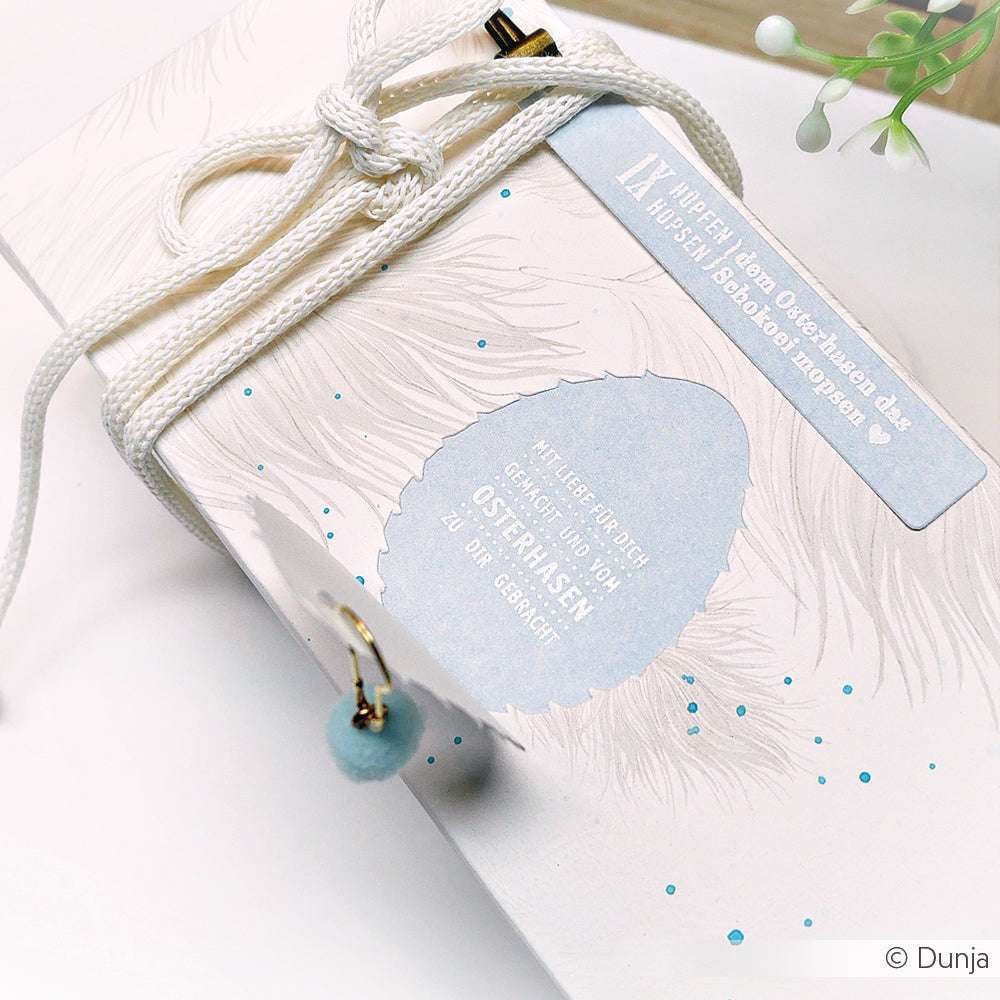 Nahaufnahme einer weißen, österlich gestalteten Geschenkbox mit blauen Sprenkeln, einem Federmotiv, das mit einem Alexandra Renke Clear Stamp gestempelt wurde, und einem dekorativen Anhänger.