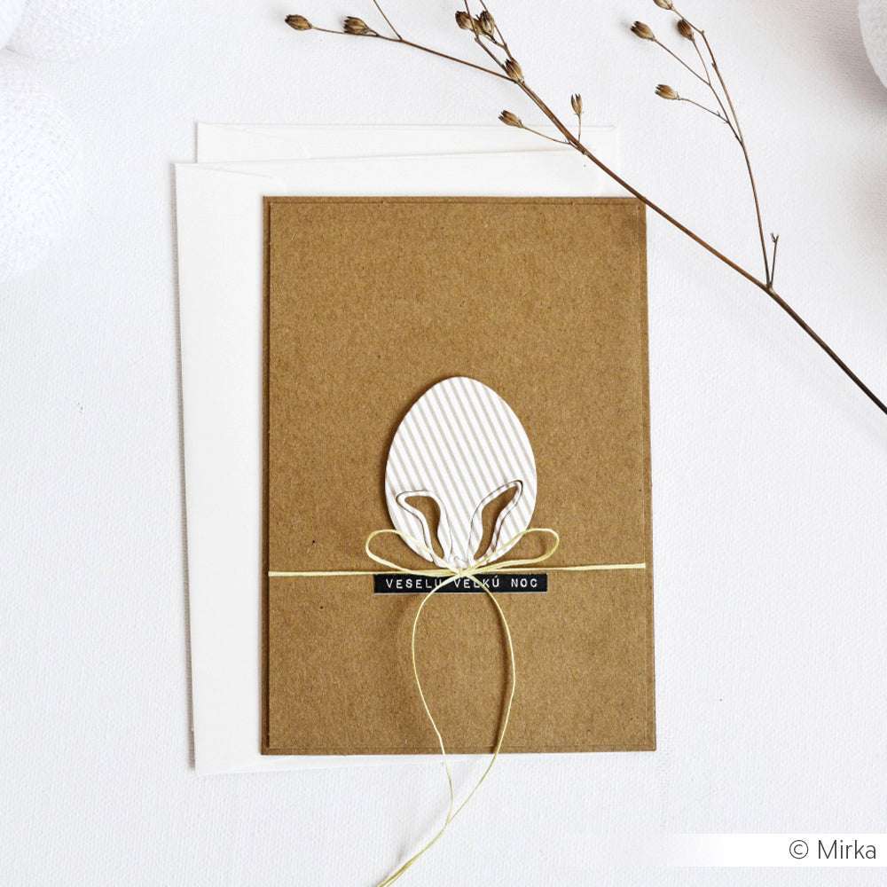 Eine handgefertigte Karte von Alexandra Renke, hergestellt mit DieCut Systemen und der Motivstanze Ei mit Hasenohren, zeigt ein dekoratives Ei und eine Schleife auf braunem Papier, eingefasst in einen weißen Umschlag mit getrockneten Zweigen für rustikalen Charme.