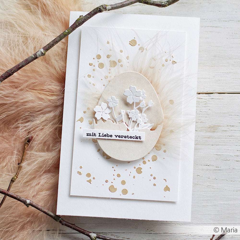 Handgefertigte Karte mit Designpapier von Alexandra Renke, einem mit der Eierstanze gestanzten Ei aus Papier, weißen Blumen, deutschem Text auf weichen Federn und zarten Farbspritzern.