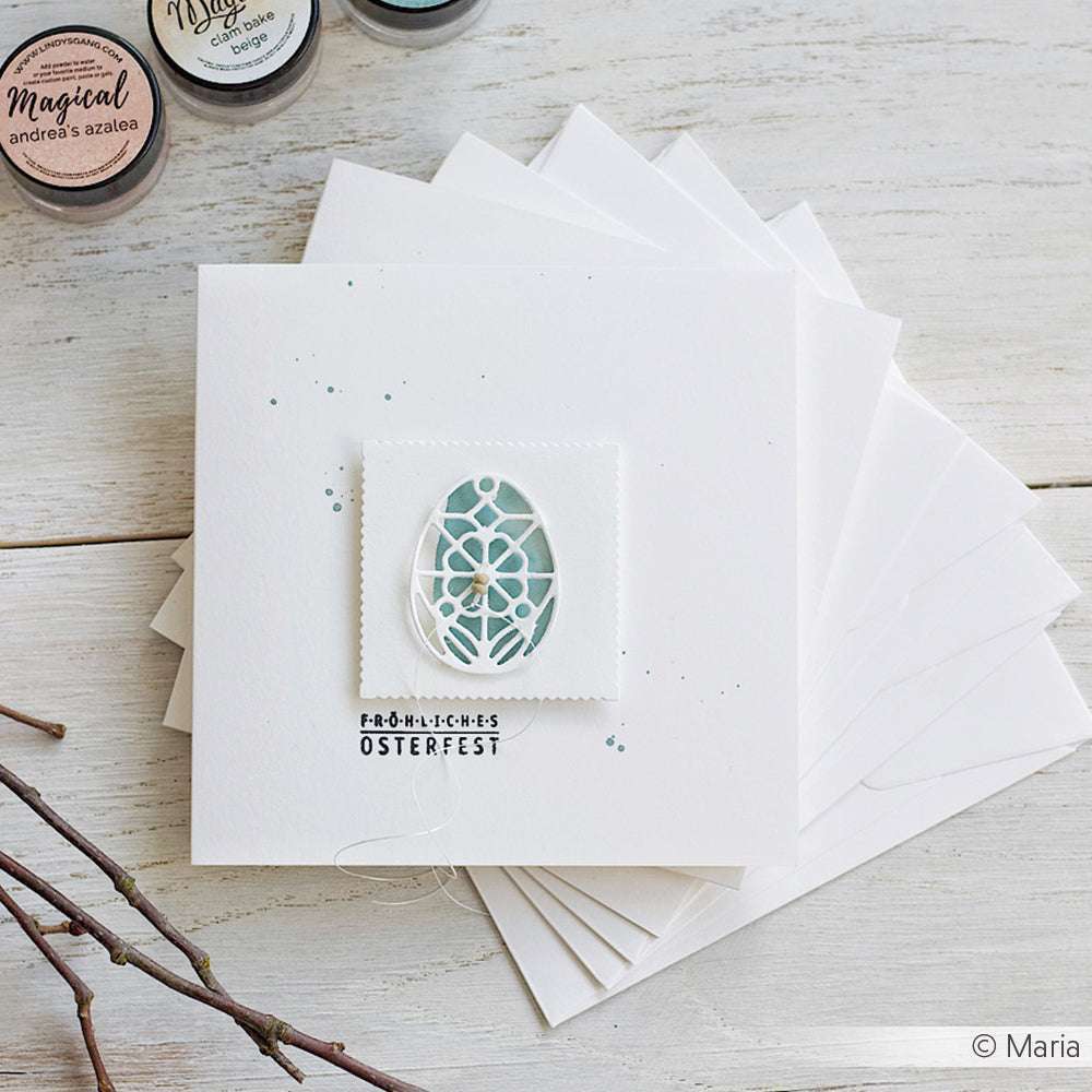 Eine handgefertigte Osterkarte von Alexandra Renke mit einem floralen Eierausschnitt von DieCut Systeme und Designpapieren ruht auf gestapelten weißen Umschlägen auf einem rustikalen Tisch.