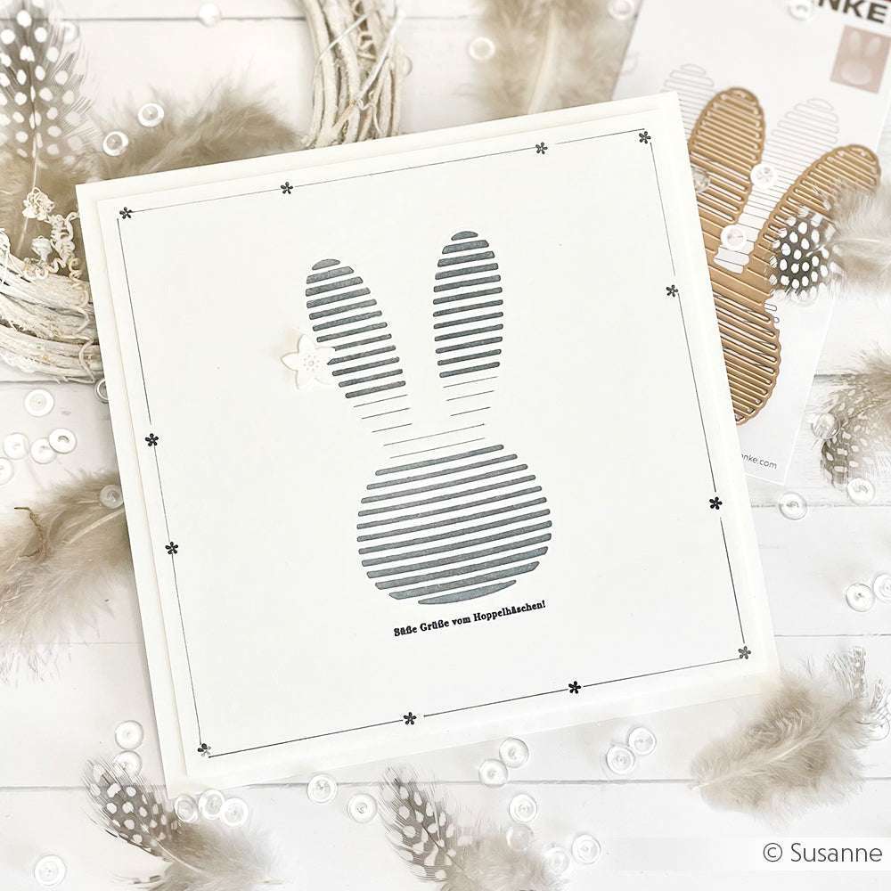 Minimalistische, quadratische Osterkarte mit einer gestreiften Hasensilhouette, die mit der Motivstanze Lamellen Hasenkopf, deutschem Text und Akzenten wie Federn und Knöpfen gestaltet wurde - ein charmantes Design von Alexandra Renke.