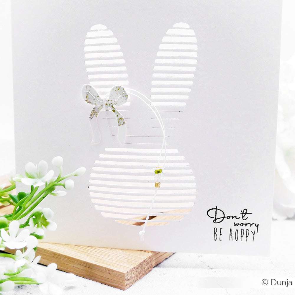 Weiße Karte mit einem ausgeschnittenen Hasen, der mit der Alexandra Renke Motivstanze Lamellen Hasenkopf hergestellt wurde, verziert mit einer Schleife, Perlen und dem Text "Don't worry, BE HOPPY". Blumen in der Nähe. Teil der bezaubernden PAPPcut®ies-Kollektion.