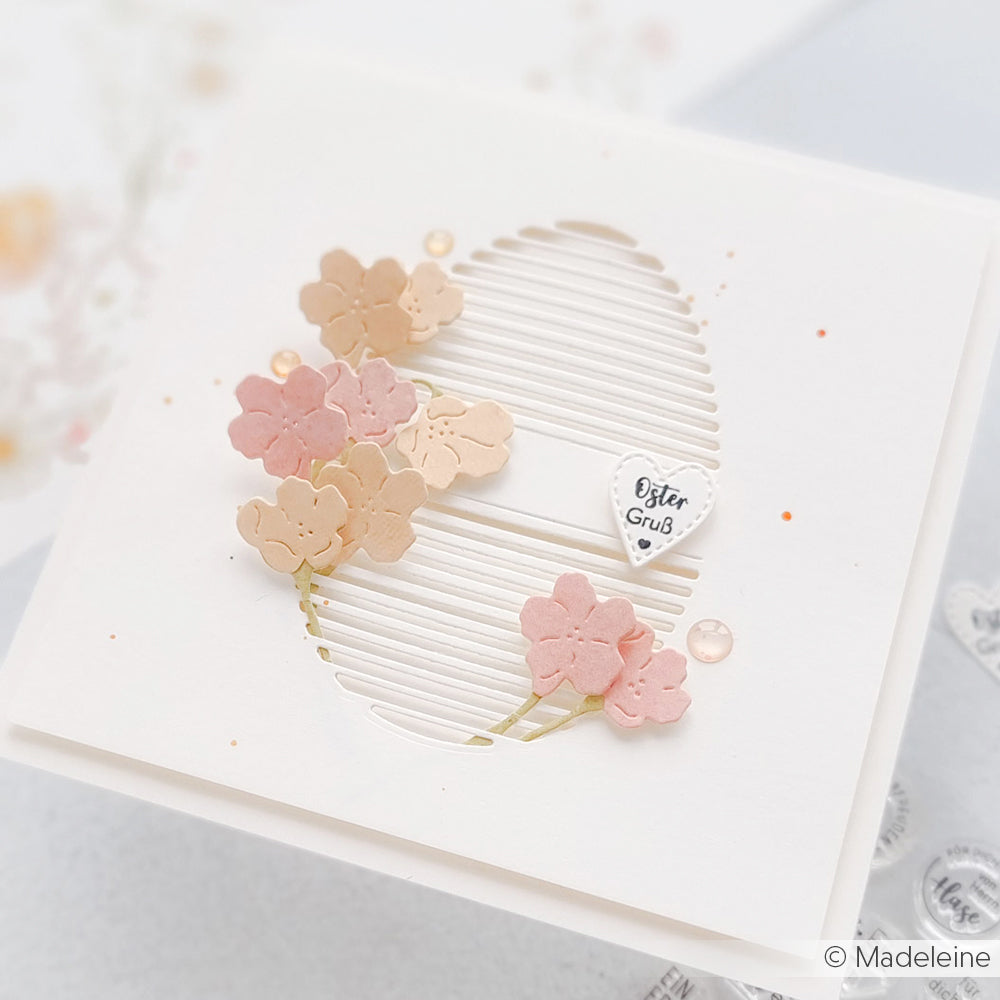 Eine weiße Karte von Alexandra Renke mit einem Motivstanze Lamellen Ei Ausschnitt, verziert mit rosa Blumen und einem kleinen Herz mit den Worten "Oster Gruß.