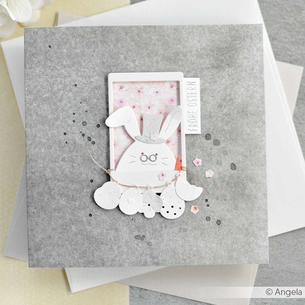 Handgemachte Osterkarte von Alexandra Renke mit einem Hasen, pastellfarbenen Blumen und dem Text "Frohe Ostern" auf grauem Hintergrund, hergestellt mit PAPPcut®ies und Deep Edge Motivstanzen für zusätzliche Details.