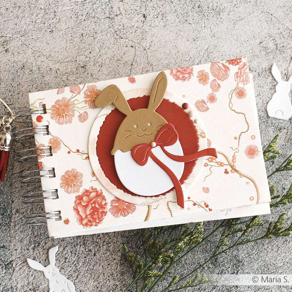 Ein dekoratives Notizbuch von Alexandra Renke mit einem Hasenmotiv und roten Blumenakzenten auf dem Einband, platziert auf einer strukturierten Oberfläche - ideal für Fans der Kollektionen Motivstanze Ei mit Hasenhüten oder PAPPcut®ies.