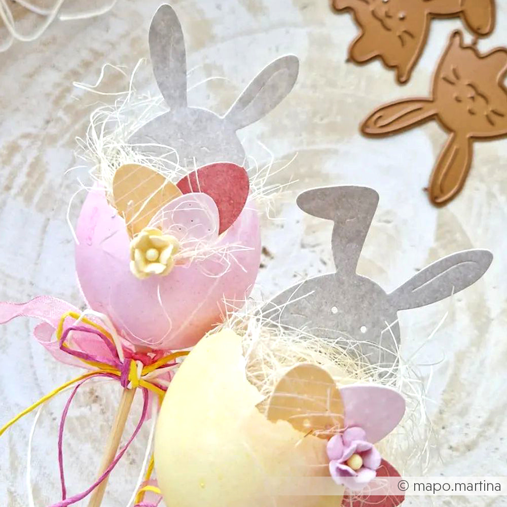 Dekorierte Eier mit Hasenohren aus Papier, die mit der Alexandra Renke Ei mit Hasenhüten Stanze gemacht wurden, pastellfarbene Herzen und Blumen von Deep Edge Motivstanzen, alles mit bunten Bändern auf einer hellen Fläche gebunden.