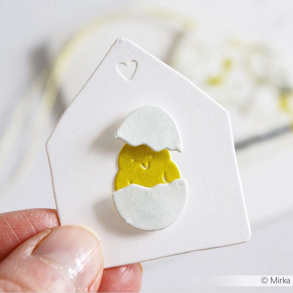Eine Hand hält eine weiße Karte mit einem lächelnden gelben Küken, das aus einer Eierschale schlüpft. Die Stanzformen von Alexandra Renke sorgen für reizvolle saisonale Details.