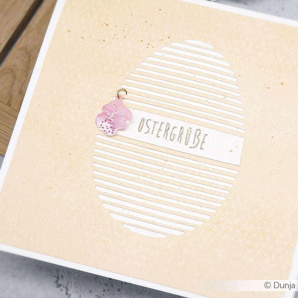 Quadratische Karte von Alexandra Renke mit einem gestreiften Ei aus Deep Edge Motivstanzen, dem Text "OSTERGRÜßE" und einem kleinen rosa Hasenanhänger auf beigem Hintergrund.