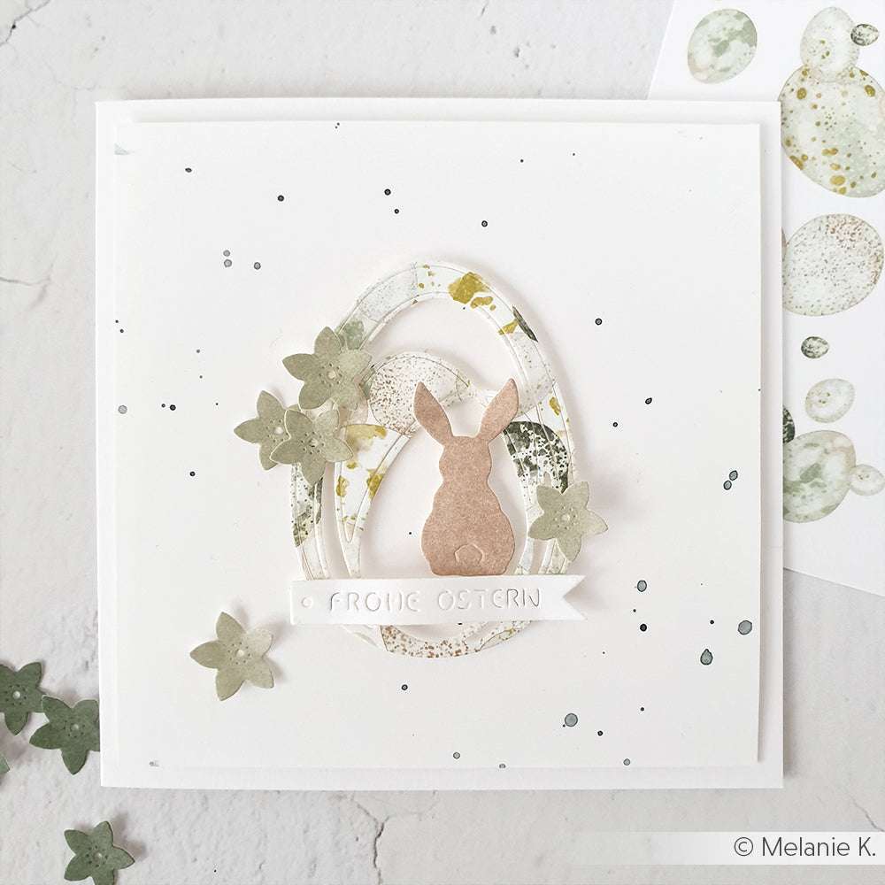 Osterkarte von Alexandra Renke mit einer Hasensilhouette, einem ovalen Rahmen mit Motivstanze Ei groß, grünen Blumen und dem Text "Frohe Ostern" auf weißem Hintergrund.