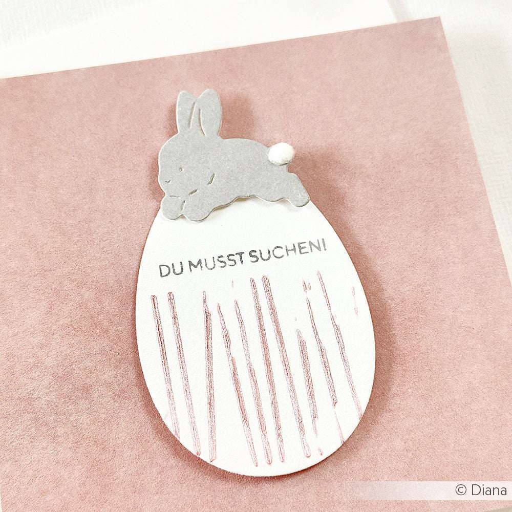 Ein grauer Papierhase, der mit der Motivstanze Hase mit Osterei von Alexandra Renke gebastelt wurde, sitzt auf einer eiförmigen Karte mit der Aufschrift "DU MUSST SUCHEN!" auf rosa - perfekt für PAPPcuties-Sammler.