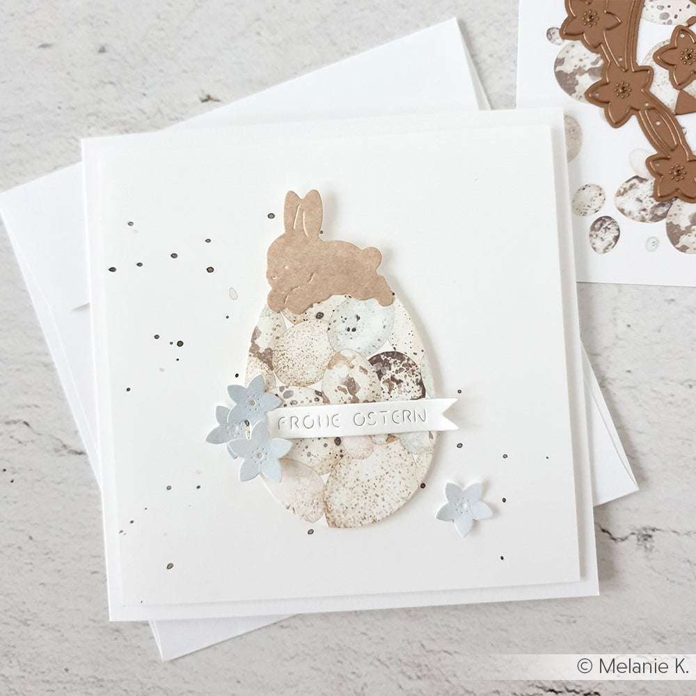 Minimalistische Osterkarte von Alexandra Renke mit einem gesprenkelten Ei, einer Hasensilhouette aus der Motivstanze Hase mit Osterei, blauen Blumen und einem "Frohe Ostern"-Banner.