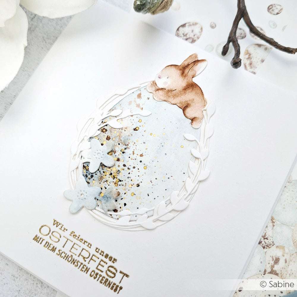Handgemachte Osterkarte von Alexandra Renke mit einem Hasen auf einem verzierten Ei, hergestellt mit Motivstanze Hase mit Osterei und PAPPcut®ies. In zartem Blau und Gold gehalten, mit deutschem Text am unteren Rand.