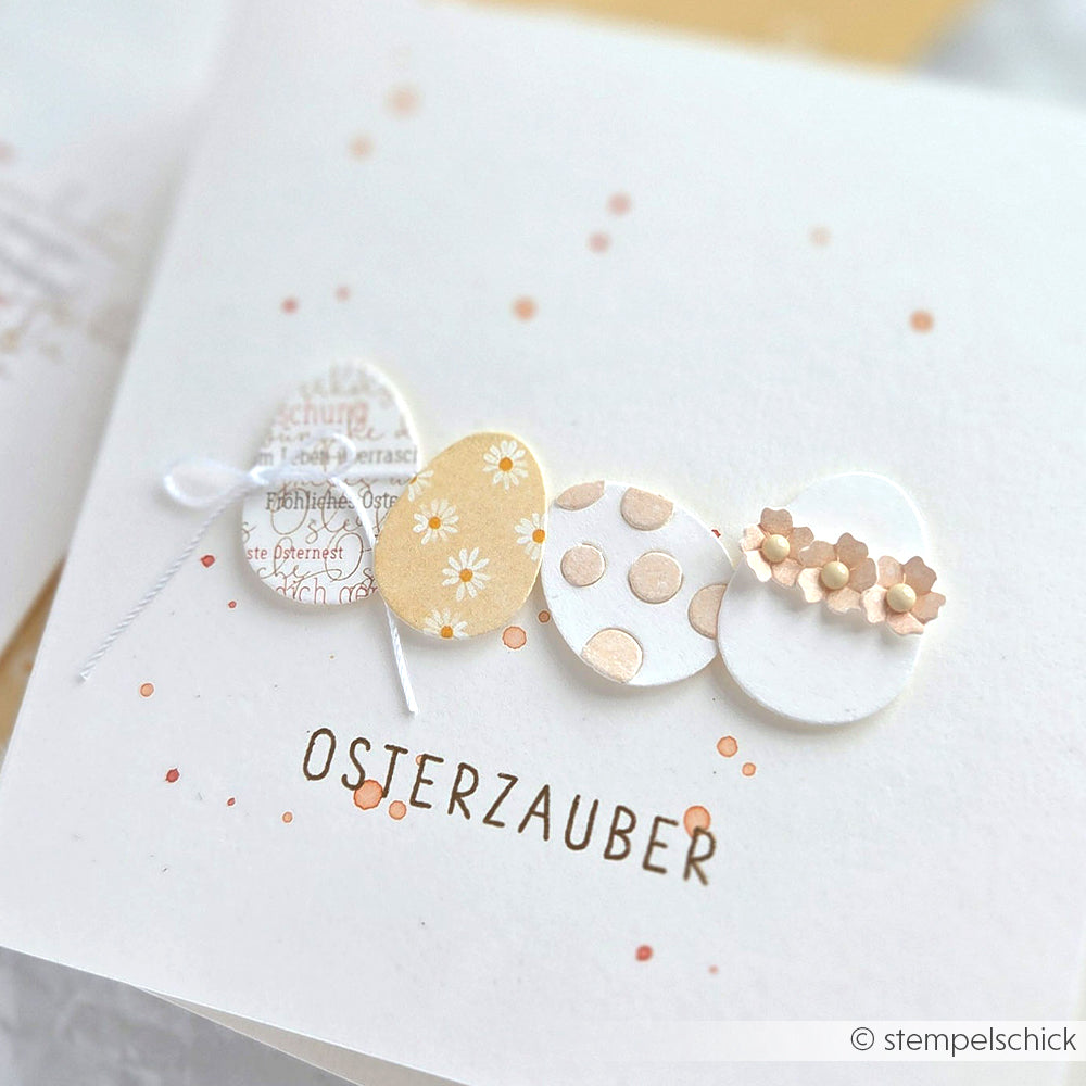 Vier verzierte Ostereier mit pastellfarbenen Mustern und Blumen über dem Wort "Osterzauber" - die perfekte Inspiration für frühlingshafte Bastelprojekte mit Alexandra Renke Designpapieren oder Ihrer Lieblingsmotivstanze.