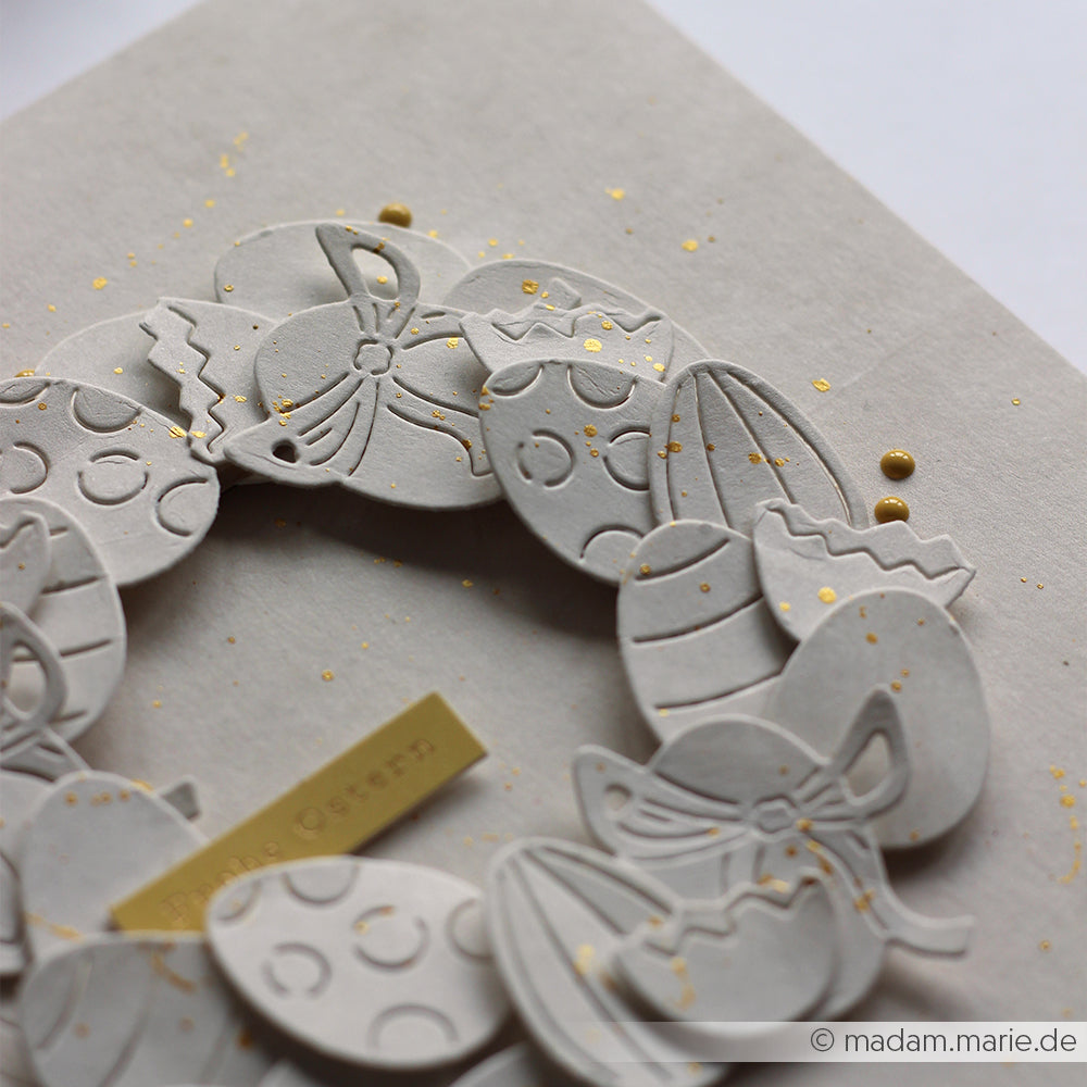 Handgefertigter Papierkranz von Alexandra Renke mit geprägten Ostereiern, hergestellt mit der Motivstanze Fröhliches Eier-Set und hochwertigen Designpapieren. Der Kranz steht auf einem hellen Hintergrund, der mit goldenen Farbspritzern akzentuiert ist.
