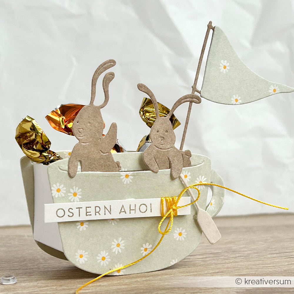 Papierhasen und eine Fahne in einer Blumentasse mit Bonbons, beschriftet mit OSTERN AHOI und einem gelben Band - gebastelt mit Alexandra Renke Motivstanze Ostern und Designpapieren für festlichen Charme.