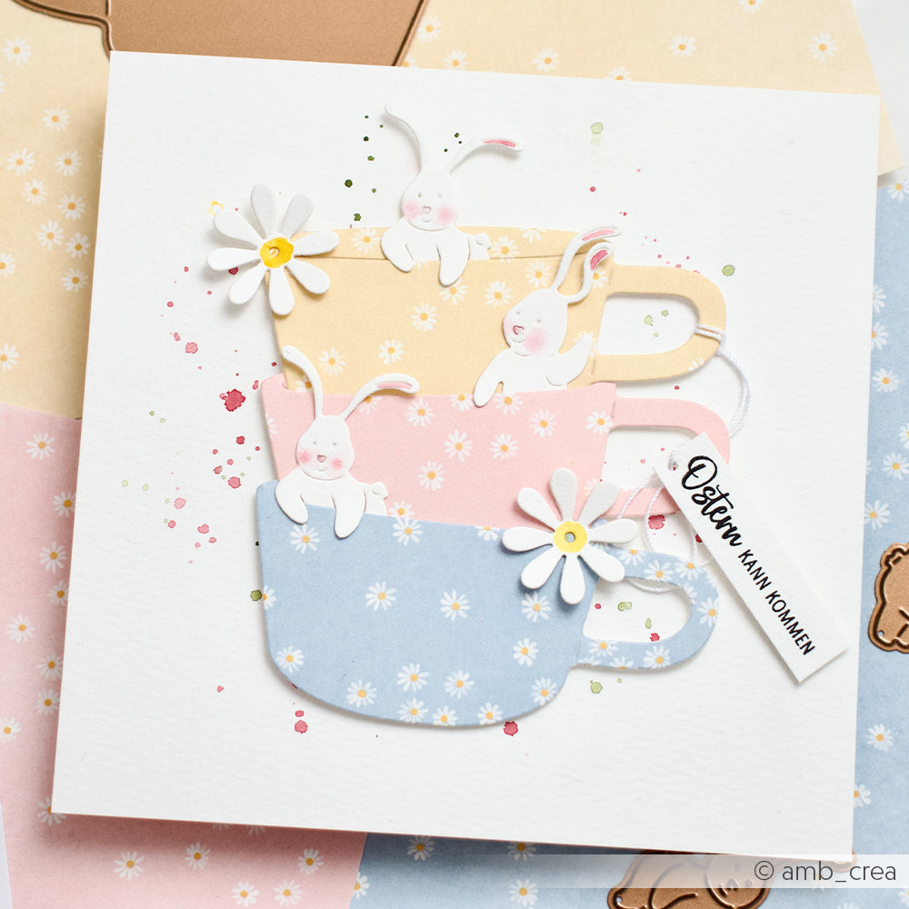 Gestapelte pastellfarbene Teetassen mit Gänseblümchen und niedlichen Hasenmotiven auf einer Blumenkarte mit einem "Beste Mama"-Anhänger, gestaltet mit Alexandra Renke Designpapieren und Motivstanze Ostern für eine besondere Note.
