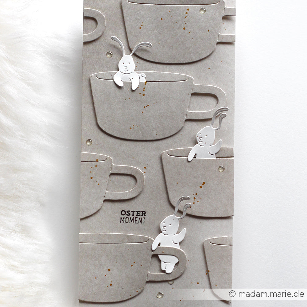 Graue Pappbecher mit weißen Hasenausschnitten, gestaltet mit Alexandra Renke Motivstanze Ostern und Designpapieren, auf einem strukturierten Hintergrund.