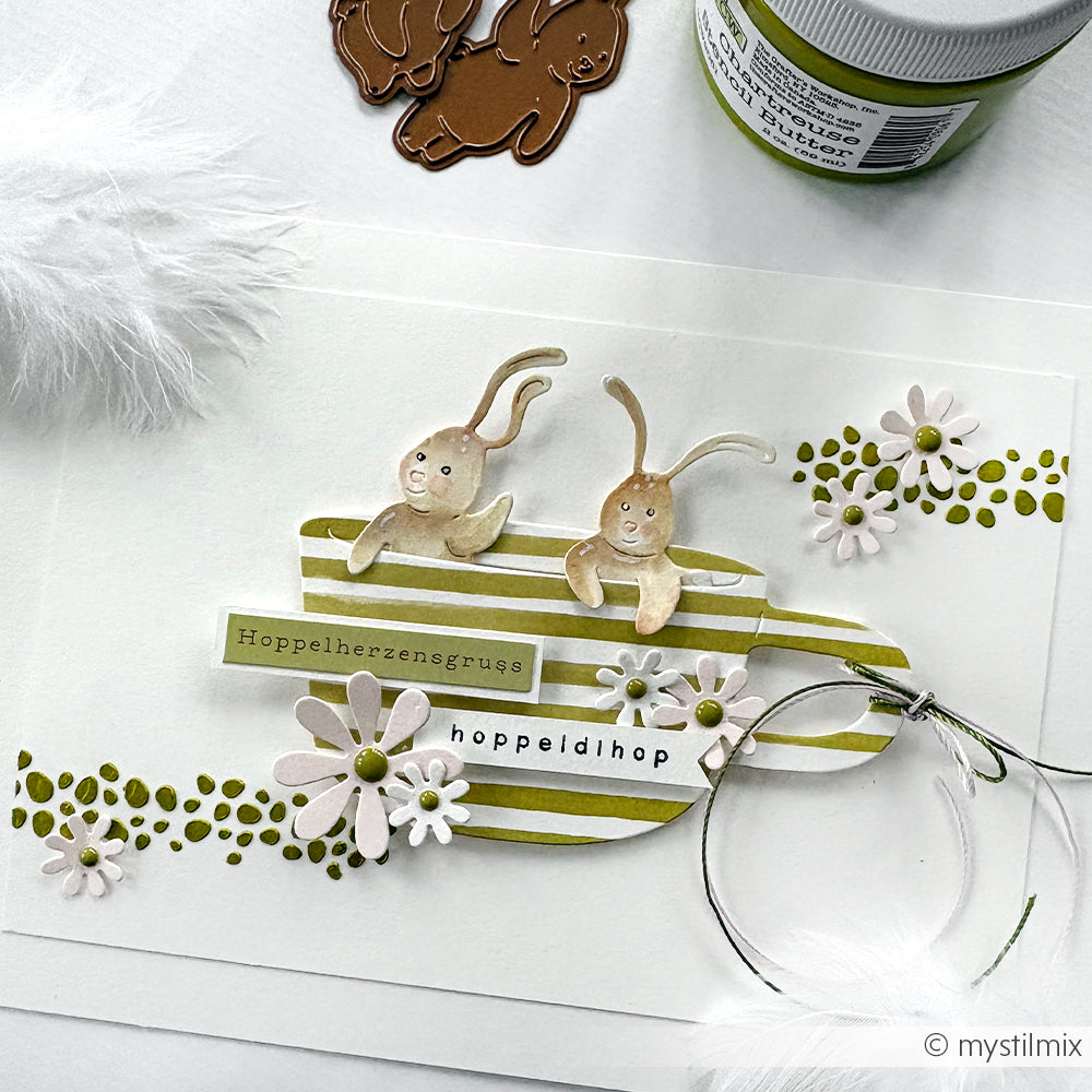 Handgemachte Karte von Alexandra Renke mit zwei Cartoon-Hasen, Blumen, Federn und deutschem Text auf weißem und grünem Hintergrund. Hergestellt mit Motivstanze Ostern und Stanzsysteme für knackige Ostermotive.