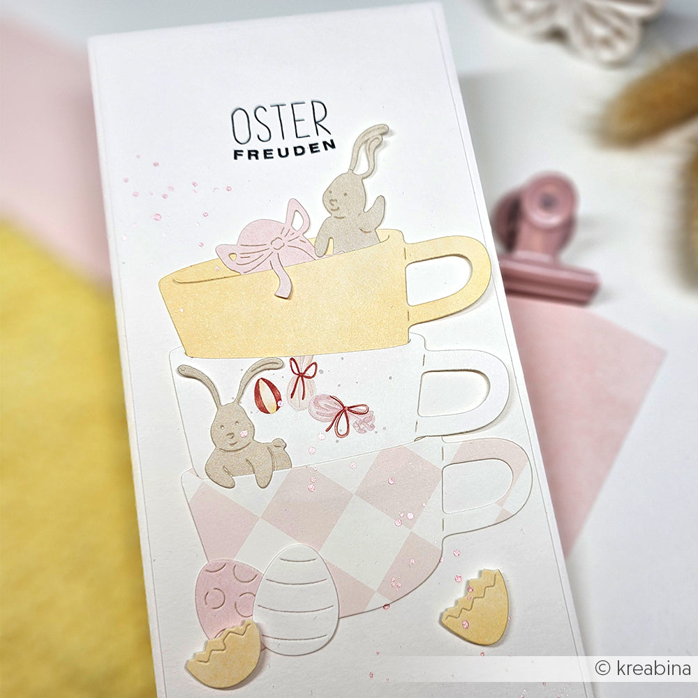 Drei pastellfarbene Teetassen übereinander gestapelt, verziert mit Papierhasen, Eiern und Verzierungen in Form von zerbrochenen Eiern, hergestellt mit Alexandra Renke Motivstanze Ostern, präsentiert auf einer strahlend weißen Karte.