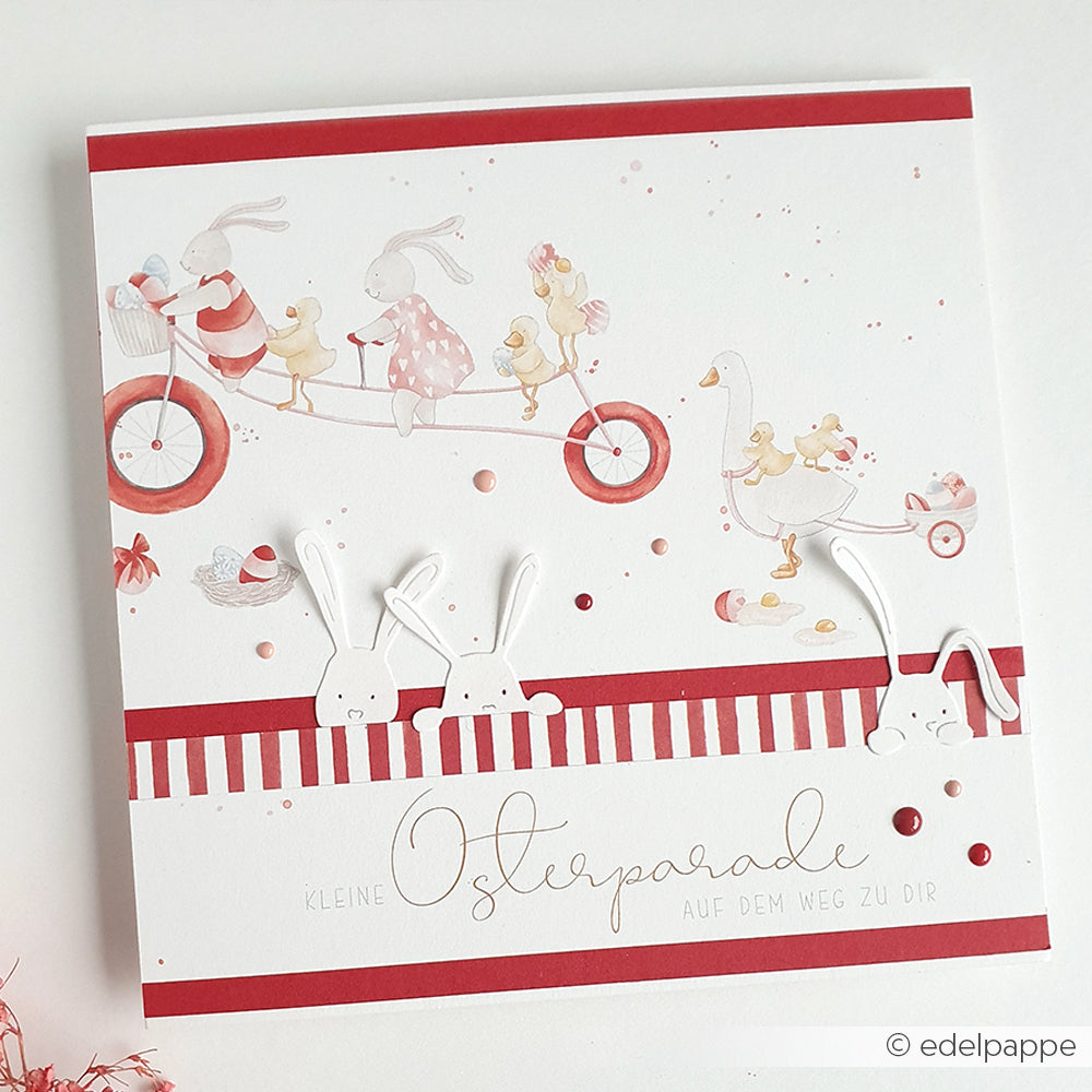 Die Osterkarte von Alexandra Renke zeigt Tiere auf einem Fahrrad und einer Parade, verziert mit roten und weißen Streifen, Hasenausschnitten und verspielten Details unter Verwendung von Designpapiere und Motivstanze Neugierige Häschen DieCut Systeme.
