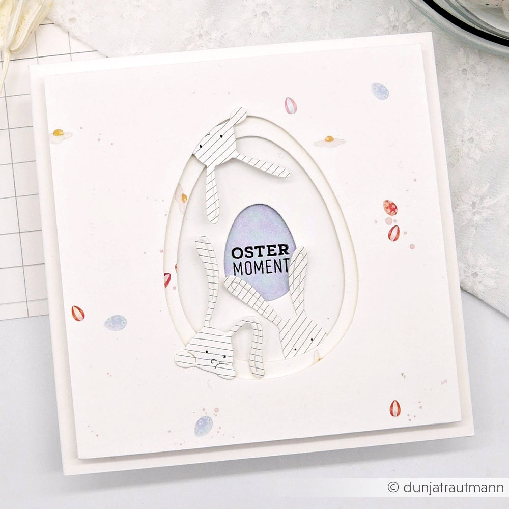Handgemachte Osterkarte mit zwei gestreiften Hasensilhouetten, die mit der Alexandra Renke Motivstanze Neugierige Häschen gestaltet wurden, sowie "Oster Moment" in einer pastellfarbenen Ei-Form.