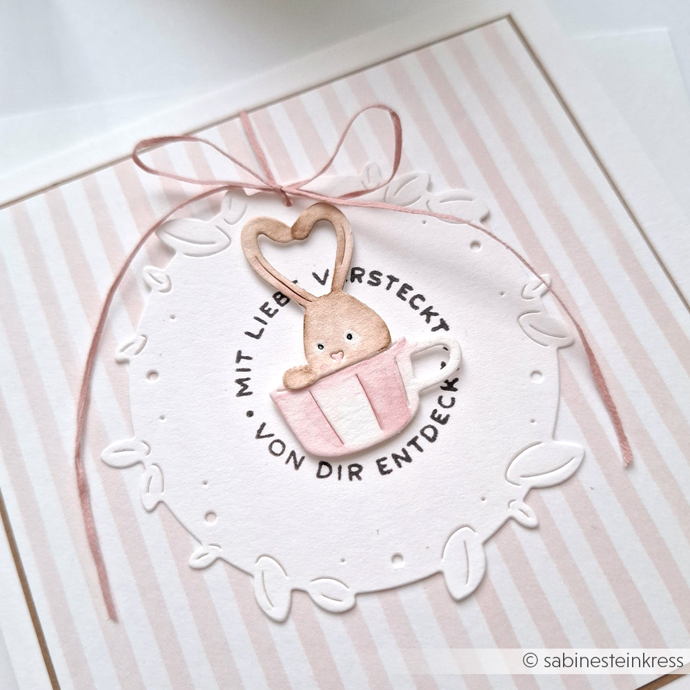 Ein niedlicher Hase in einer Teetasse mit Herz, deutschem Text und einer rosa Schleife auf Streifen - ideal für Alexandra Renke Designpapiere oder die Motivstanze Neugierige Häschen Stanzsysteme.