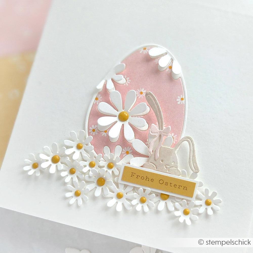 Osterkarte mit Gänseblümchen, einem Hasen und einem rosafarbenen Ei, mit "Frohe Ostern" auf einem gelben Etikett. Verarbeitet mit Alexandra Renke DieCut Systeme und Motivstanze Osterei mit Margeriten für charmante Details.