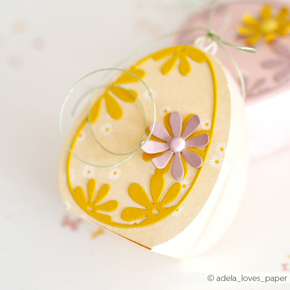 Eiförmige Papierschachtel von Alexandra Renke, verziert mit gelben und lilafarbenen Blumen und einer grünen Kordelschleife auf der Oberseite - hergestellt mit Motivstanze Osterei Dies für präzise, charmante Details.