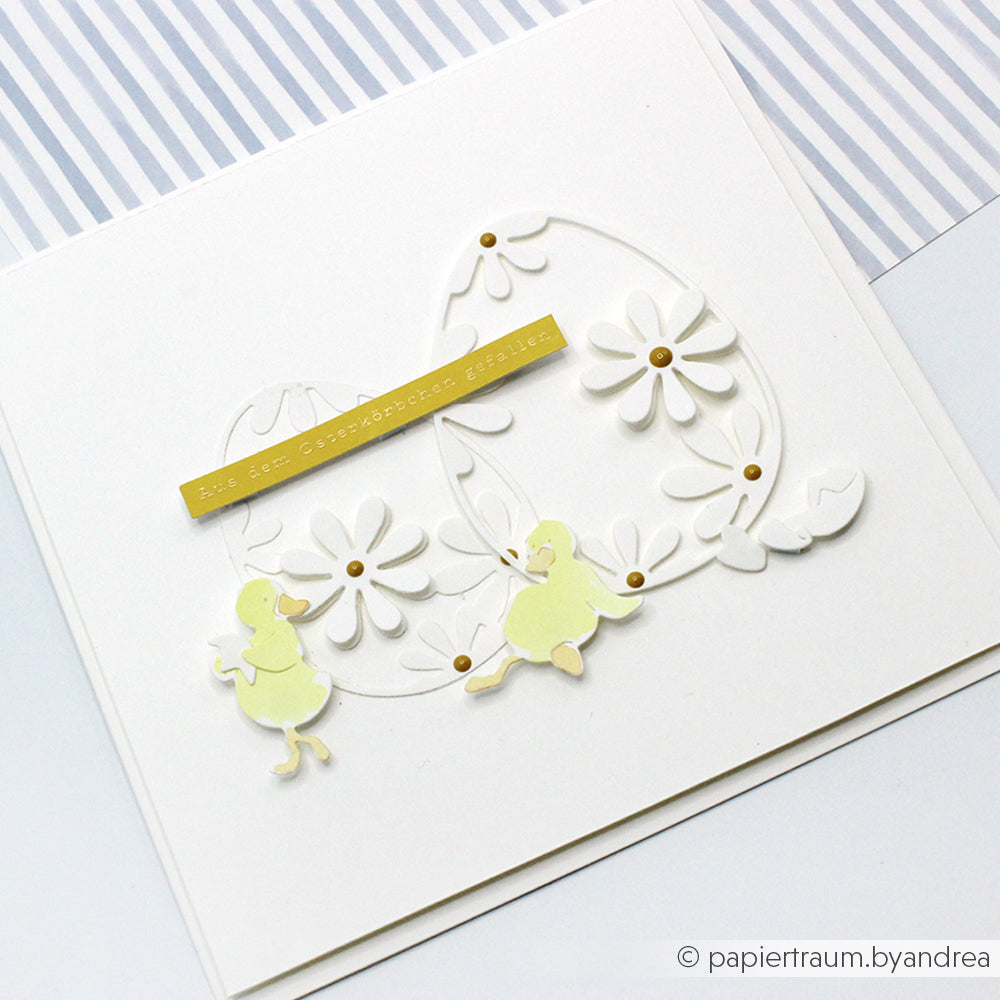 Weiße Grußkarte von Alexandra Renke mit Gänseblümchen, einer Eierkontur mit Motivstanze Osterei, gelben Küken und einem goldenen Streifen mit deutschem Text - ideal, um Ihre Lieblingsdesignpapiere hervorzuheben.