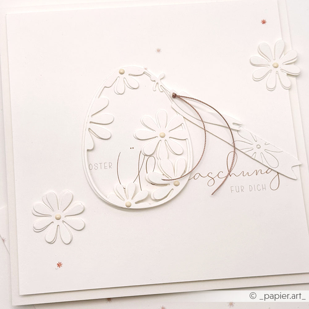 Weiße Osterkarte von Alexandra Renke mit geprägten Blumen, einem eiförmigen Ausschnitt (Motivstanze Osterei), deutschem Text und zarter brauner Schnur.