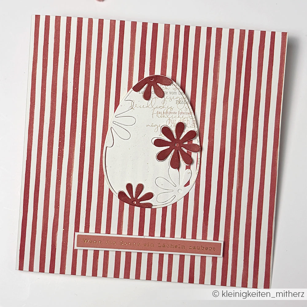 Die quadratische Karte von Alexandra Renke mit roten und weißen Streifen, hergestellt mit Stanzsystemen und Motivstanze Osterei, zeigt eine Eiform mit roten Blumen in der Mitte und einem Textetikett darunter - ideal für kreative Dies Basteln Projekte.