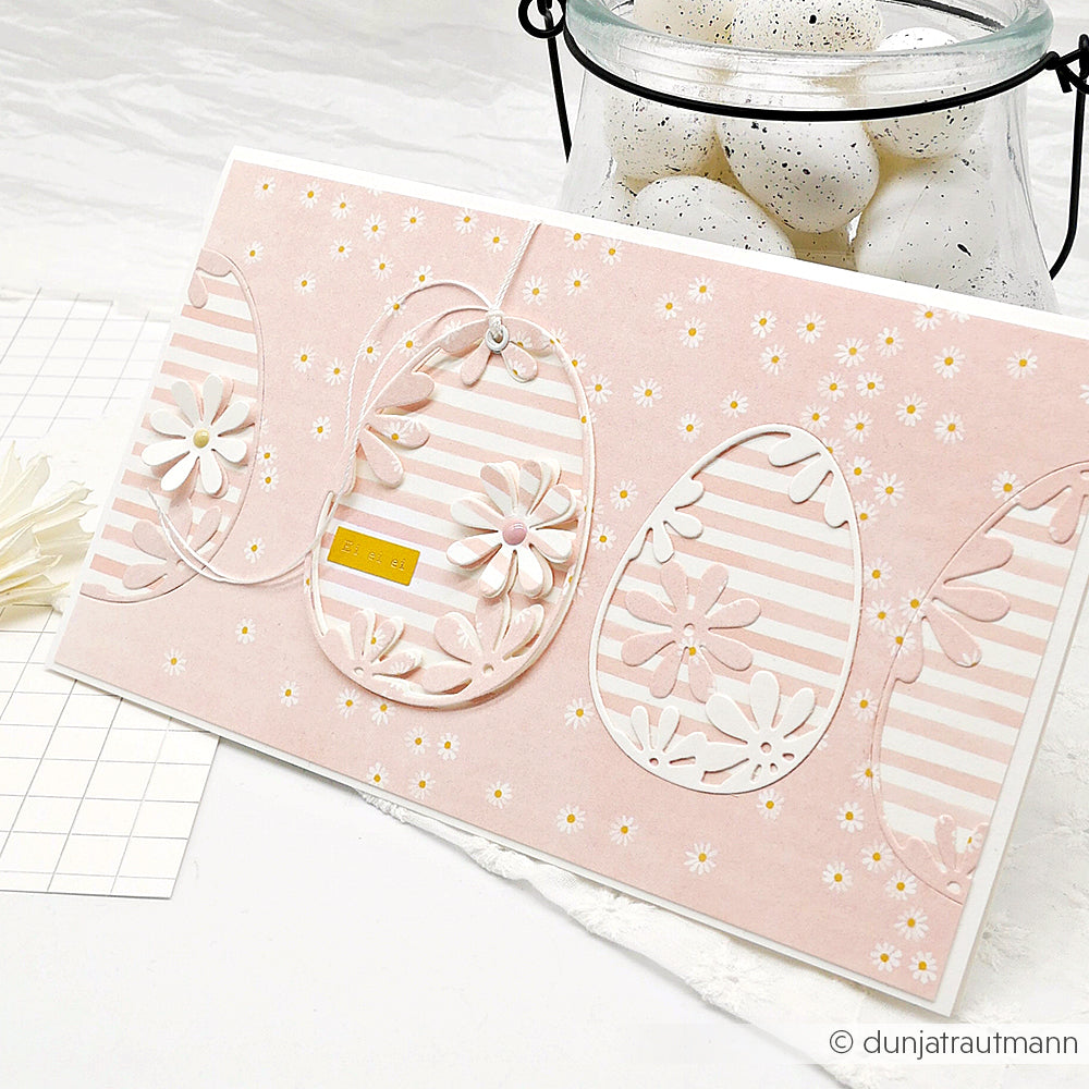 Rosafarbene Osterkarte mit floralen und gestreiften Eimotiven, hergestellt mit Alexandra Renke Dies für Basteln, wie der Motivstanze Osterei, die neben einem Glas mit gesprenkelten weißen Eiern abgebildet ist.