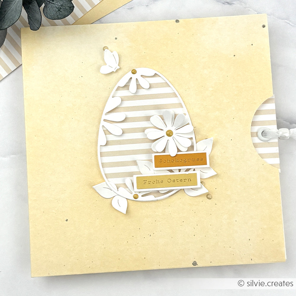 Gelbe Osterkarte von Alexandra Renke mit einem gestreiften Ei aus Motivstanzen, weißen Blumen und dem Text "Frohe Ostern" auf der Vorderseite.