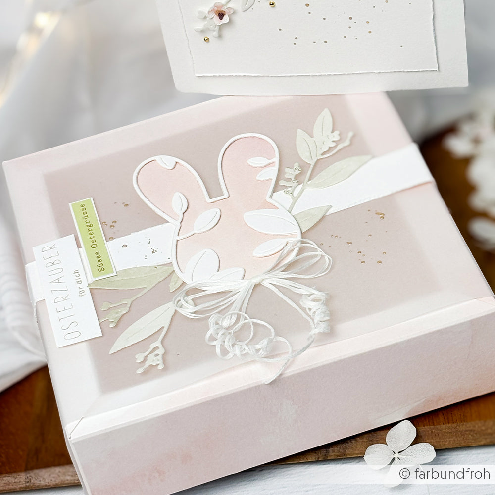 Eine pastellrosa Oster-Geschenkbox von Alexandra Renke, verziert mit einer Motivstanze Hasenkopf, einer weißen Schleife aus Garn und floralen Akzenten.