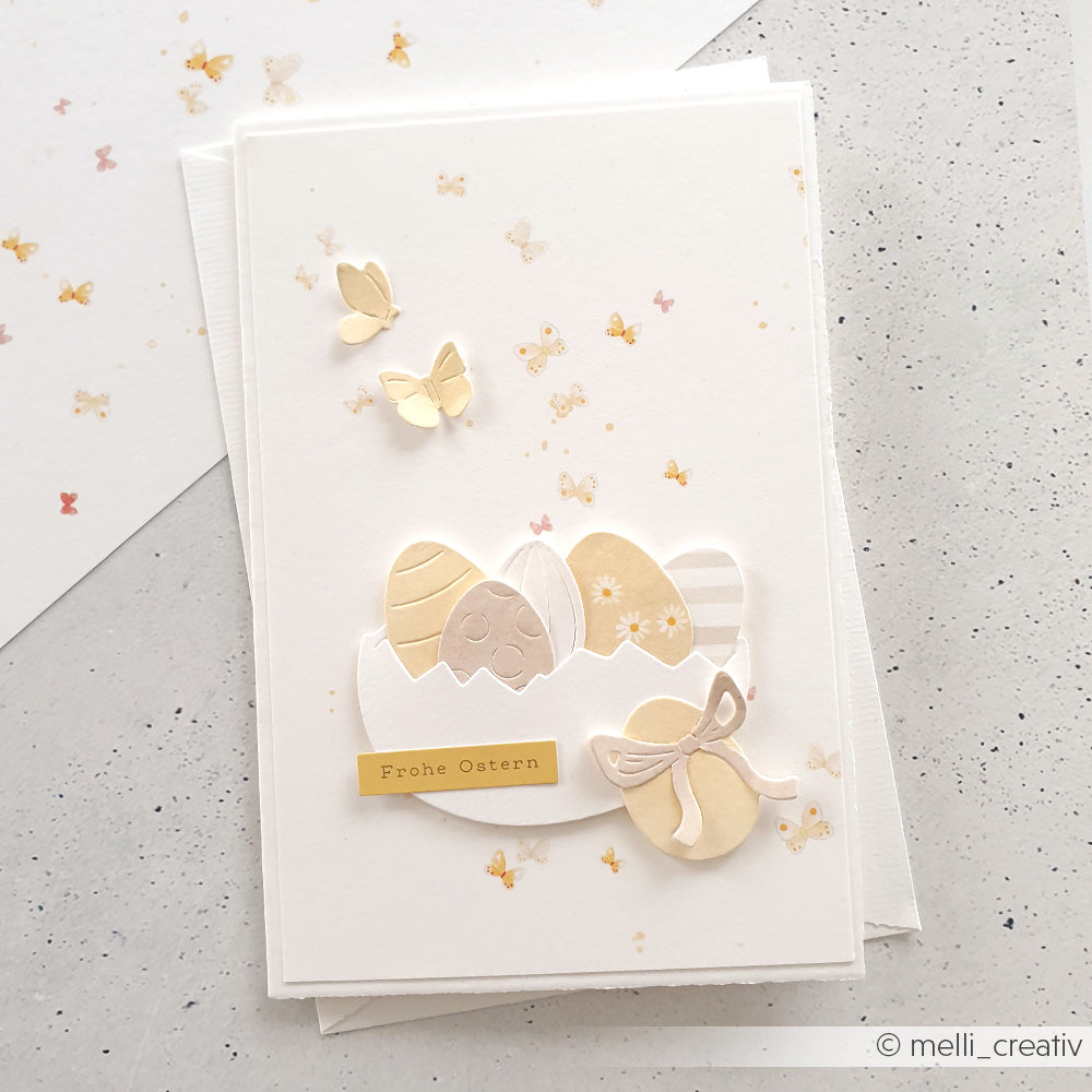 Handgemachte Osterkarte von Alexandra Renke aus Designpapier mit pastellfarbenen Eiern, Schmetterlingen und dem Text "Frohe Ostern". Gestanzte Eierhälften sorgen für charmante Details auf einem strahlend weißen Hintergrund.