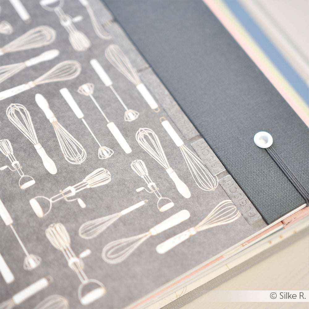 Schwarz-weißes Alexandra Renke Designpapier mit Küchenutensilien neben dunklem Stoff mit weißem Knopf - ideal für Buchbinderei oder kreative Papierprojekte.