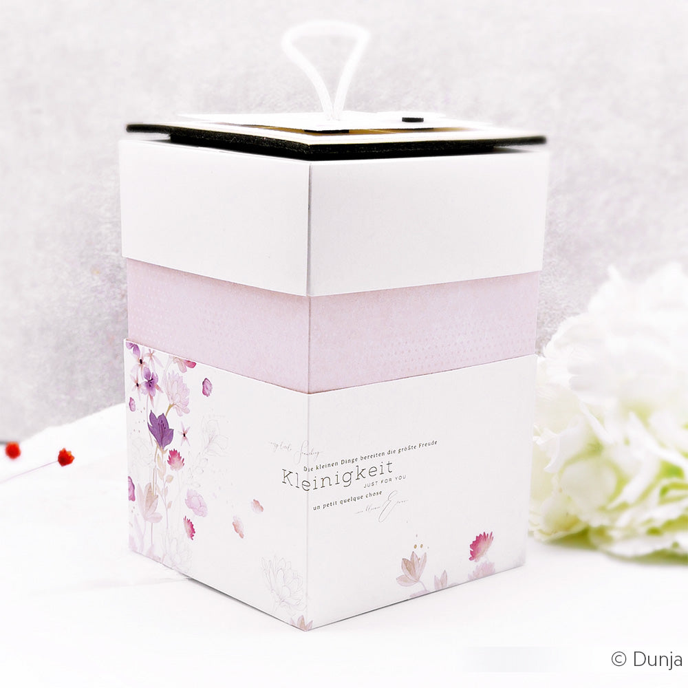 Eine pastellfarbene Blumengeschenkbox von Alexandra Renke, die mit Designpapier und einem zartrosa Band gestaltet wurde und auf einer weißen Fläche neben weißen Blumen steht.