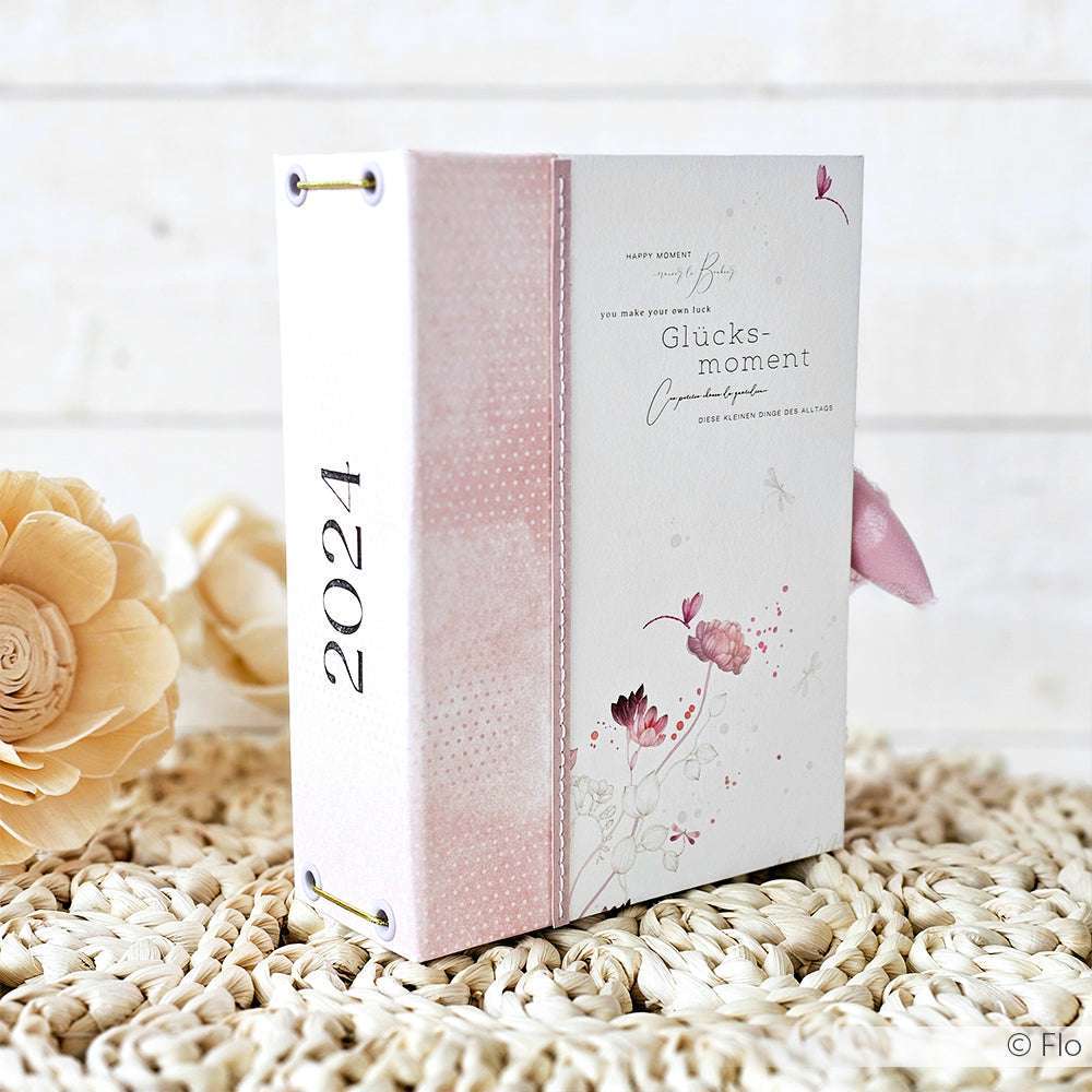 Ein floraler Planer 2024 von Alexandra Renke mit rosa Akzenten steht aufrecht auf einer gewebten Matte neben dekorativen Blumen - perfekt für Papierprojekte oder als Paar mit passenden Grußkarten.