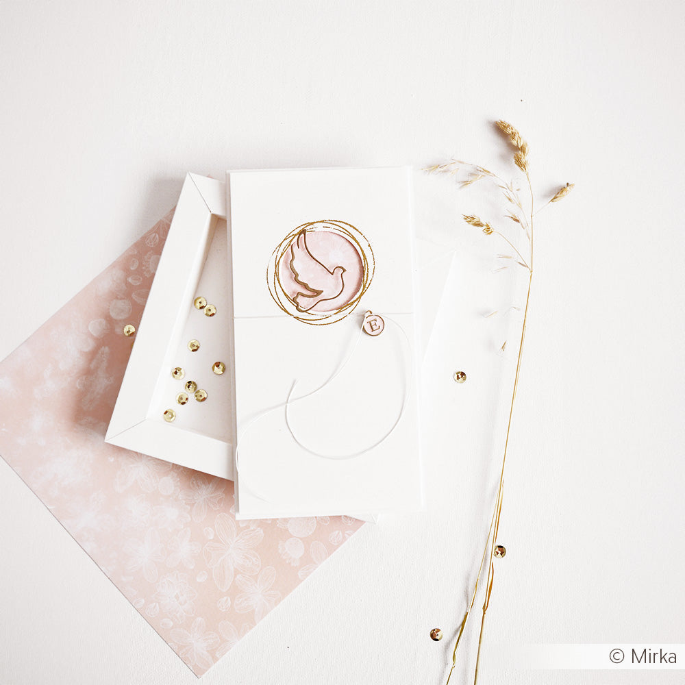 Eine weiße Karte von Alexandra Renke, die mit Designpapier gestaltet und mit einem goldenen Taubenmotiv verziert wurde, ruht auf einem rosafarbenen Umschlag inmitten von goldenen Perlen und getrocknetem Gras.