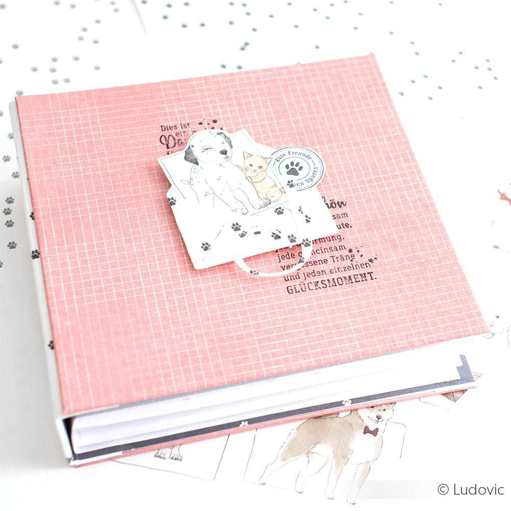 Das rosafarbene Scrapbook von Alexandra Renke, ideal für Papierprojekte, zeigt auf dem Cover eine niedliche Zeichnung von Hund und Katze. Es ist aus Designpapier gefertigt und ruht auf einer weißen Oberfläche, die mit verspielten Pfotenabdrücken verziert ist.