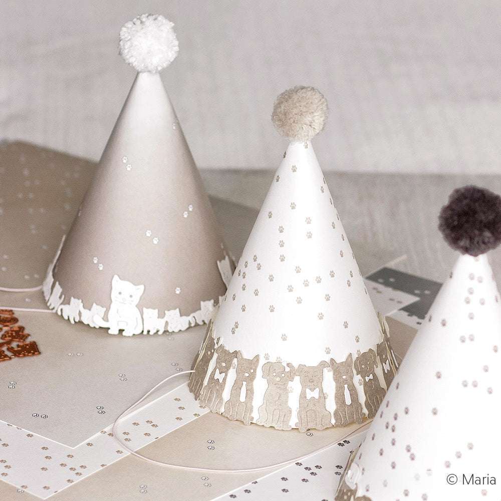 Partyhüte mit Bommeln und niedlichen Tiermotiven liegen auf einem Tisch, der mit gemustertem Designpapier von Alexandra Renke dekoriert ist.