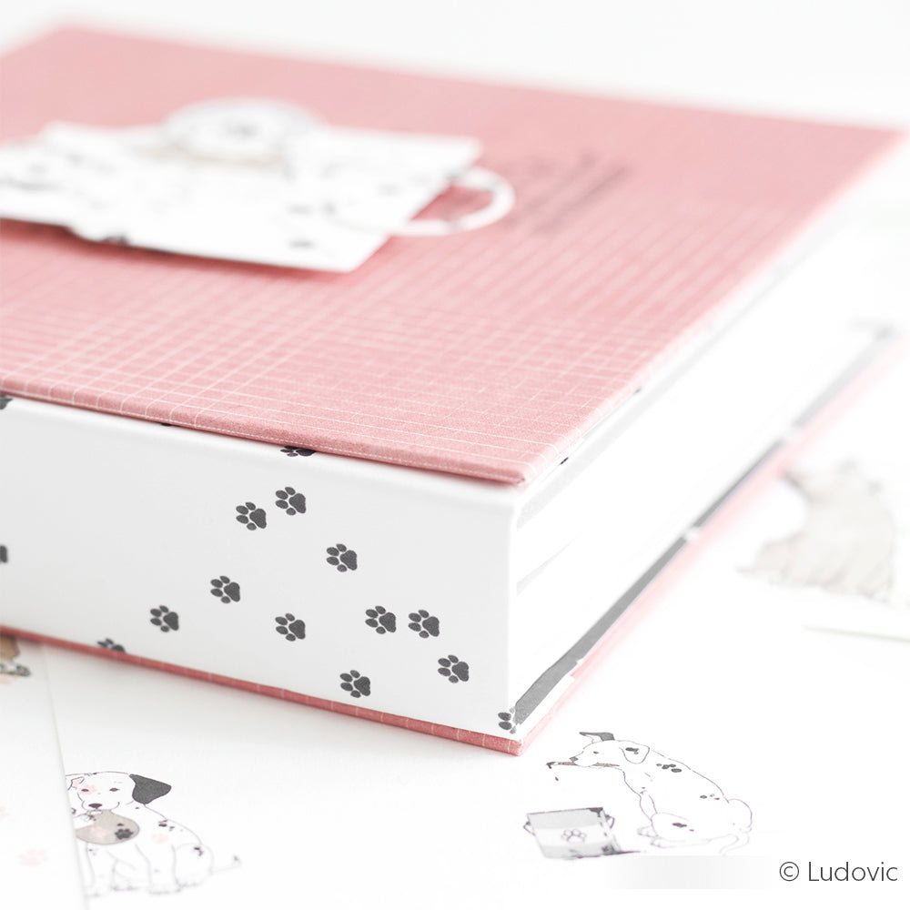 Ein rosafarbenes Buch mit schwarzen Pfotenabdrücken am Rand, umgeben von Papieren und Anhängern mit Hundemotiven - ideal zum Buchbinden oder Papierbasteln mit dem Designpapier Pfötchen Dunkelgrau von Alexandra Renke.