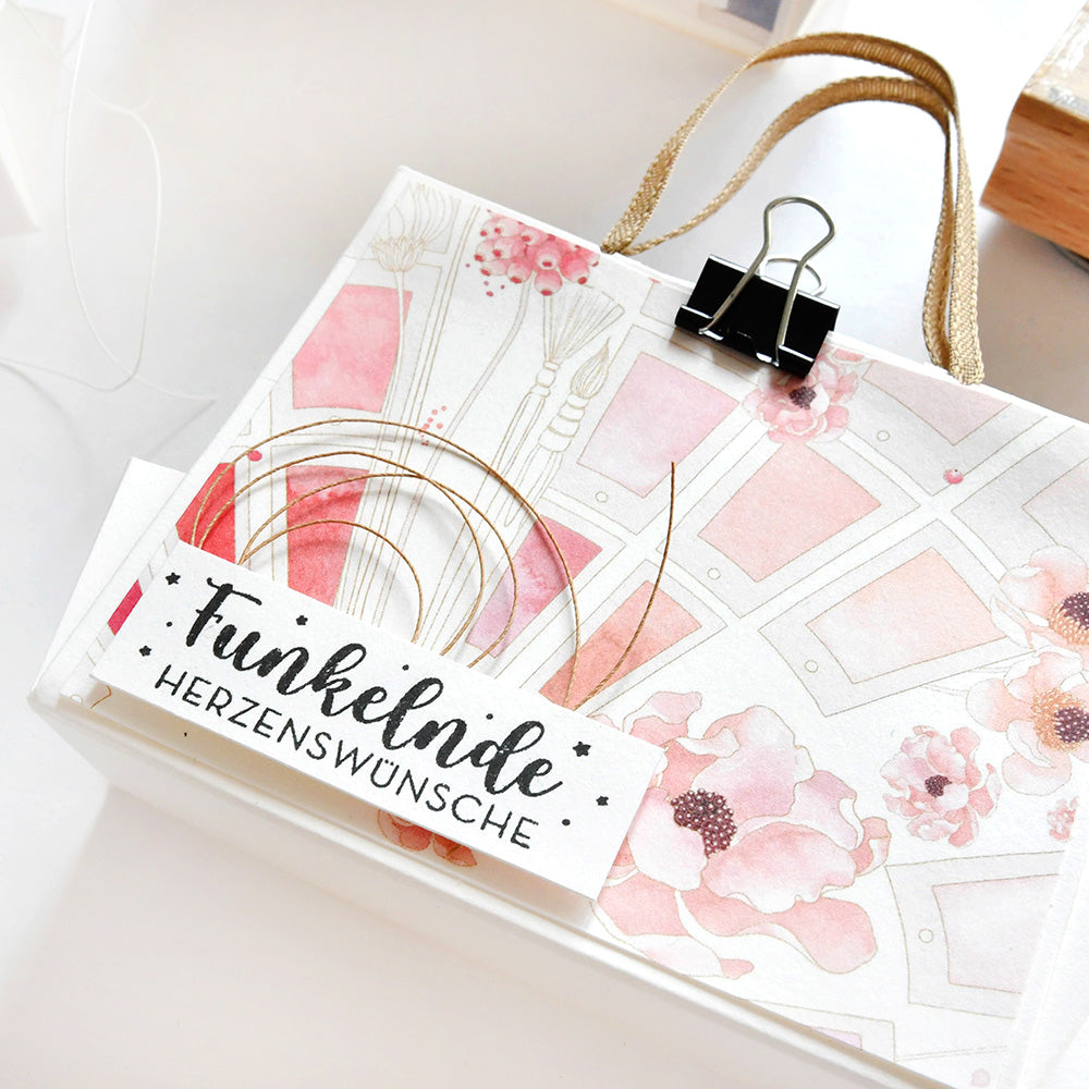 Eine floral gemusterte Geschenktasche von Alexandra Renke aus Designpapier, die oben mit einem weißen Etikett "Funkelnde Herzenswünsche" verschlossen wird - perfekt für die Präsentation von Kreative Blütenpracht.