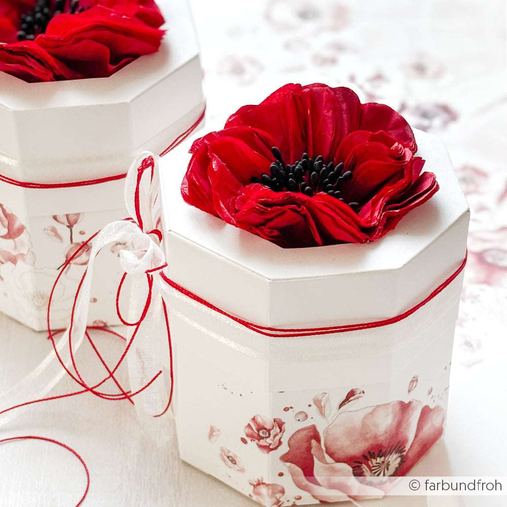 Die weiße, achteckige Geschenkbox von Alexandra Renke mit rotem Blumendeckel, Blütenpracht, floralem Designpapier und einem leuchtenden Farbkarten-Rot-Band ruht auf einer hellen Fläche.