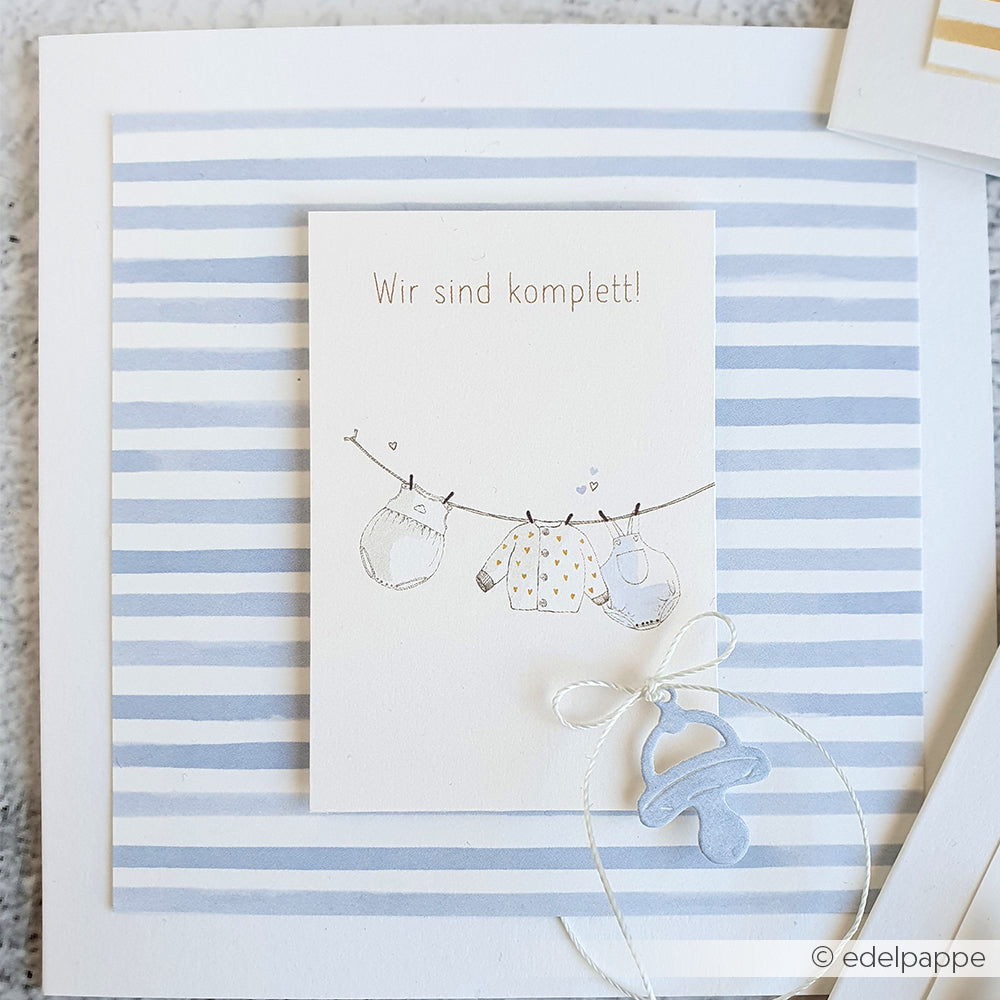 Grußkarte von Alexandra Renke mit "Wir sind komplett!" über Babykleidung auf einer Wäscheleine, verziert mit einem blauen Schnuller - ideal für Papierprojekte und Fans von stylischem Designpapier.