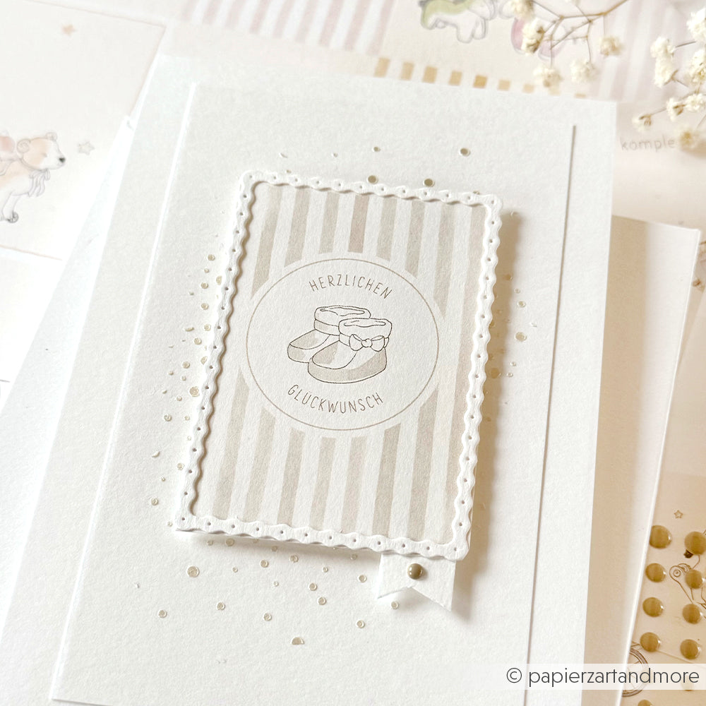 Grußkarte von Alexandra Renke mit einer Illustration von Babyschuhen, deutschem Text und einem gepunkteten Rand auf Weiß - ideal für Papierprojekte oder als Inspiration für Designpapier in der Buchbinderei.