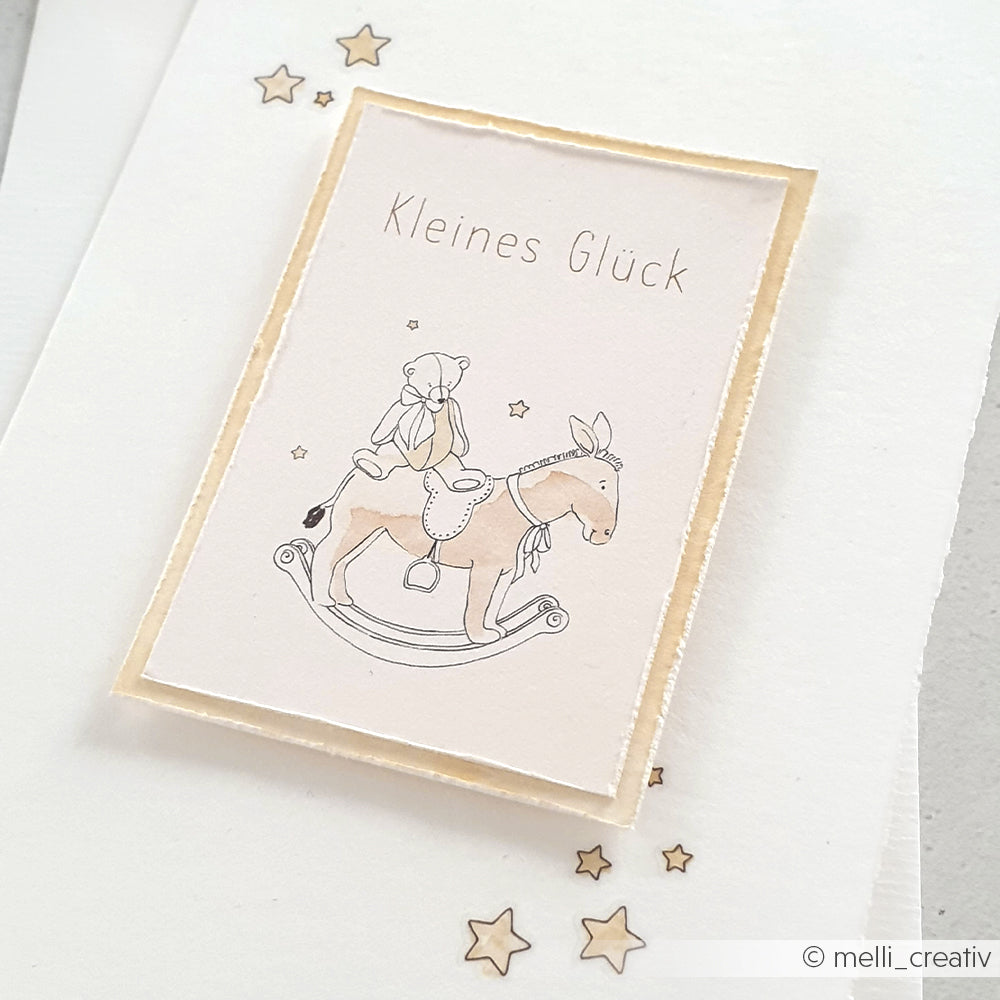 Ein niedliches Grußkarten-Motiv von Alexandra Renke: Ein Bär auf einem Schaukelpferd, umgeben von Sternen und dem Schriftzug "Kleines Glück". Ideal für Papierprojekte mit Designpapier.