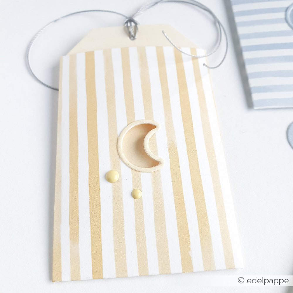 Gestreifter Anhänger von Alexandra Renke aus Designpapier mit einem Halbmondausschnitt, zwei gelben Punkten und einer silbernen Schnur auf hellem Hintergrund.